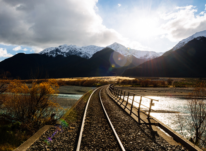 Image of New Zealand scenery, rail track in foreground going over water and towards hills in the background.